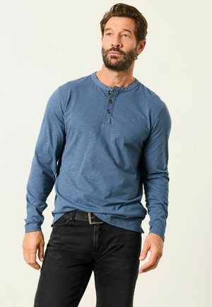 Mand med skæg iført blå langærmet henley-shirt og sorte bukser, stående mod en ensartet lys baggrund, kigger svagt mod højre.