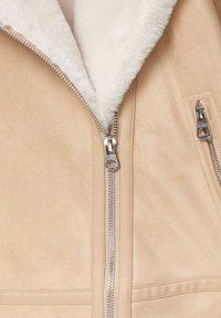 Veste en daim beige avec col en fausse fourrure blanche et fermetures éclair en métal argenté, comprenant une fermeture éclair de poche verticale et une diagonale.