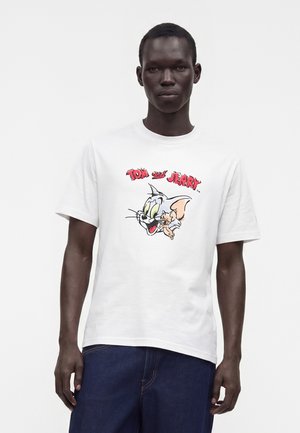 Mand iført hvid T-shirt med farverig Tom og Jerry tegneserie grafik og rød tekst, kombineret med mørkeblå bukser.