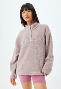 Lyserød fleece-pullover med høj krave, tre knaplukninger og struktureret overflade. Bæres med lyserøde shorts.