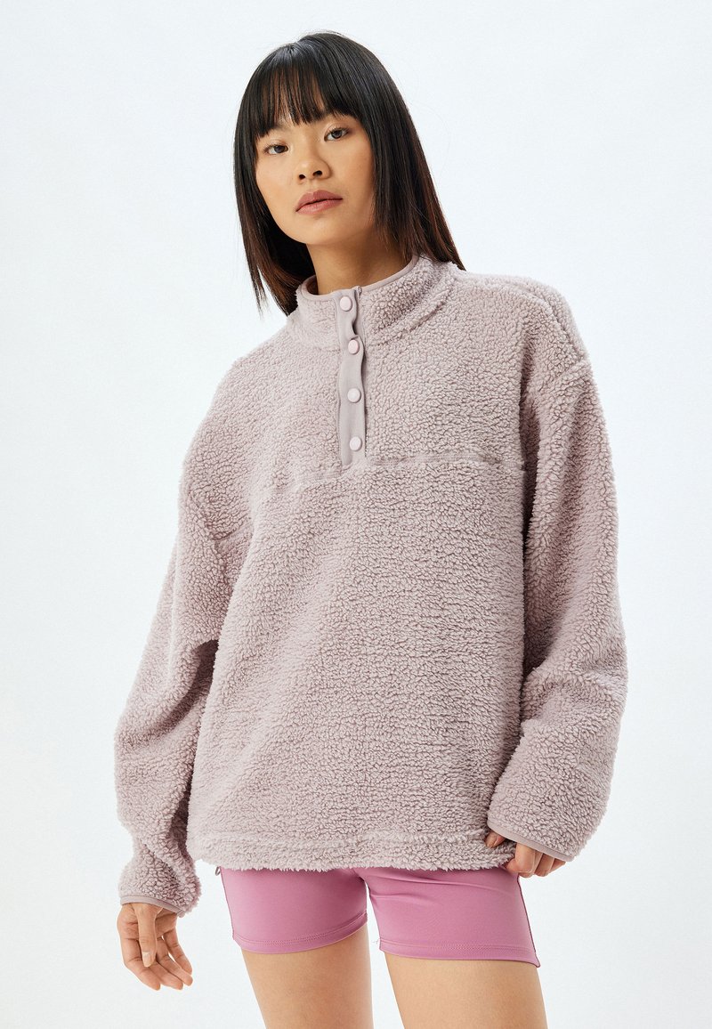 Lila Fleece-Pullover mit hochgeschlossenem Kragen, dreiknöpfiger Knopfleiste und strukturierten Oberfläche. Getragen mit pinkfarbenen Shorts.