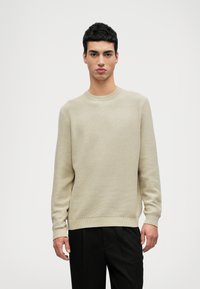 Beiger strukturiertes Strickoberteil mit Rundhalsausschnitt, langen Ärmeln und gerippten Bündchen, kombiniert mit schwarzen Hosen. Glatte Verarbeitung und lässige Passform.