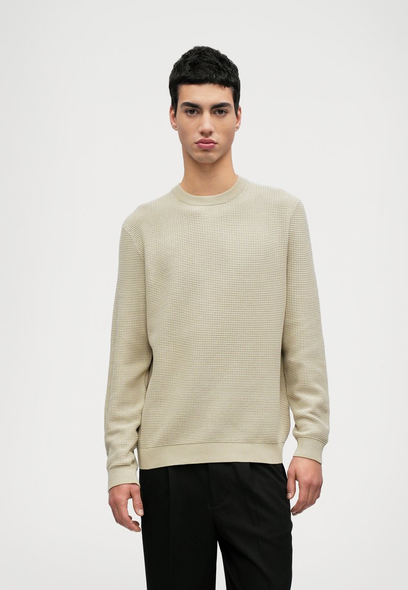 Beiger strukturiertes Strickoberteil mit Rundhalsausschnitt, langen Ärmeln und gerippten Bündchen, kombiniert mit schwarzen Hosen. Glatte Verarbeitung und lässige Passform.