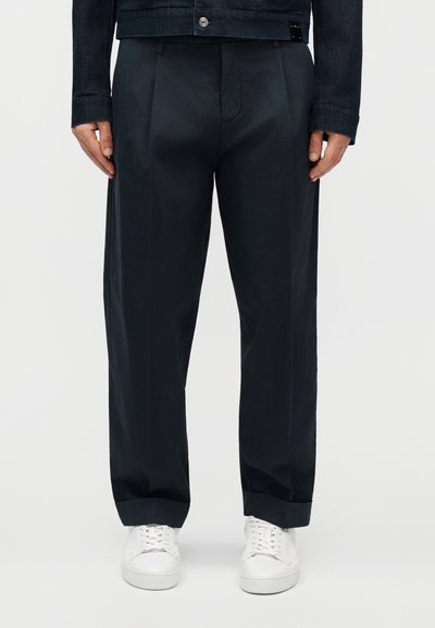 Homme portant un pantalon ajusté bleu marine foncé avec revers et des baskets blanches, mains détendues le long du corps, sur un fond uni.