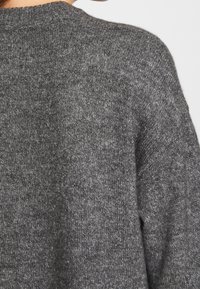 Gros plan d'une personne portant un pull en maille douce et texturée gris foncé, montrant l'épaule et le haut du dos.