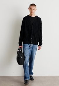 Mustat kabelineulepaidat, joissa on napit, päällä musta paita, yhdistettyinä sinisiin farkkuihin ja mustiin kenkiin. Mukana tumma laukku.