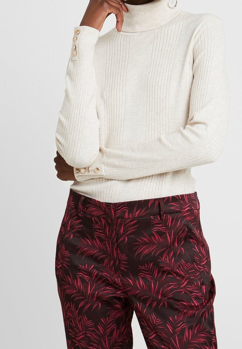 Krémový pletený rolák s ribovanou texturou a knoflíkovými detaily na rukávech, kombinovaný s tmavými kalhotami s červeným listovým vzorem.