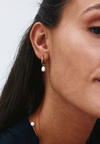 Pendiente de aro dorado con un pequeño colgante de perla blanca, que presenta una textura suave y una forma redondeada. Llevado en el lóbulo contra cabello oscuro.