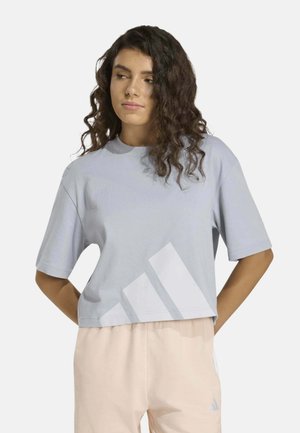Mujer joven con cabello rizado lleva una camiseta corta azul claro con un gran logo blanco de Adidas y pantalones beige claros, de pie con las manos detrás de la espalda.