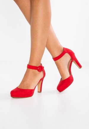 Escarpins rouges en suède avec une sangle de cheville dotée d'une boucle circulaire, bout rond et un talon beige solide. Texture lisse et design élégant.