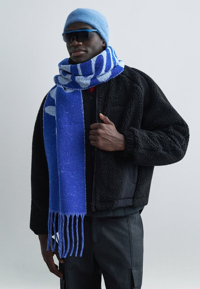 Μπλε μαντήλι με σχέδια και φράντζες, φορεμένο πάνω από μαύρο φλις μπουφάν. Σε συνδυασμό με ανοιχτό μπλε σκουφάκι και σκουρόχρωμα γυαλιά ηλίου.