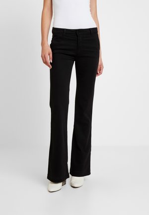 Kvinde iført sorte bootcut bukser, hvid ærmeløs top og hvide hælede sko, stående mod en ensfarvet lys baggrund.
