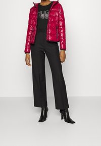Rote glänzende Puffjacke mit Kapuze, kombiniert mit einem schwarzen T-Shirt mit silbernem Grafikdesign und hochtaillierten schwarzen Hosen sowie Stiefeletten mit Absatz.