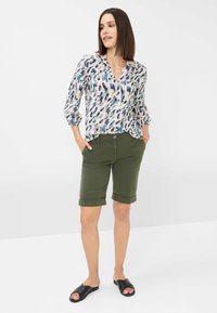 Blusa bianca con un motivo astratto blu e verde, colletto; pantaloni corti verdi con risvolto; sandali neri a scivolo.