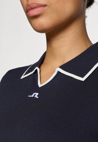 Jūros mėlynumo polo marškiniai su balta apvadu, turintys v-kaklą ir mažą logotipo užrašą ant krūtinės. Lygi megztinė tekstūra.