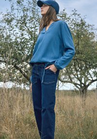 Junge Frau, die einen blauen Jeanshut, ein langärmeliges Hemd und Jeans trägt, steht auf einer grasbewachsenen Wiese mit Bäumen im Hintergrund.