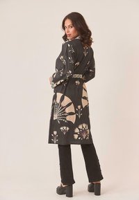 Mujer con cabello castaño ondulado vistiendo un abrigo largo negro con un patrón floral, pantalones negros y tacones de plataforma negros, posando frente a un fondo liso.