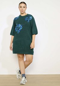 Gebreide jurk in donkergroen met korte mouwen en blauwe sequin bloemenpatronen. Losse pasvorm, knie-lengte, gecombineerd met grijze sneakers en patroon sokken.