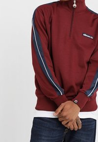 Persoon in een kastanjebruine longsleeve half-zip sweatshirt met marineblauwe en witte strepen op de mouwen, handen voor zich ineengeschoven, zichtbare blauwe jeans.