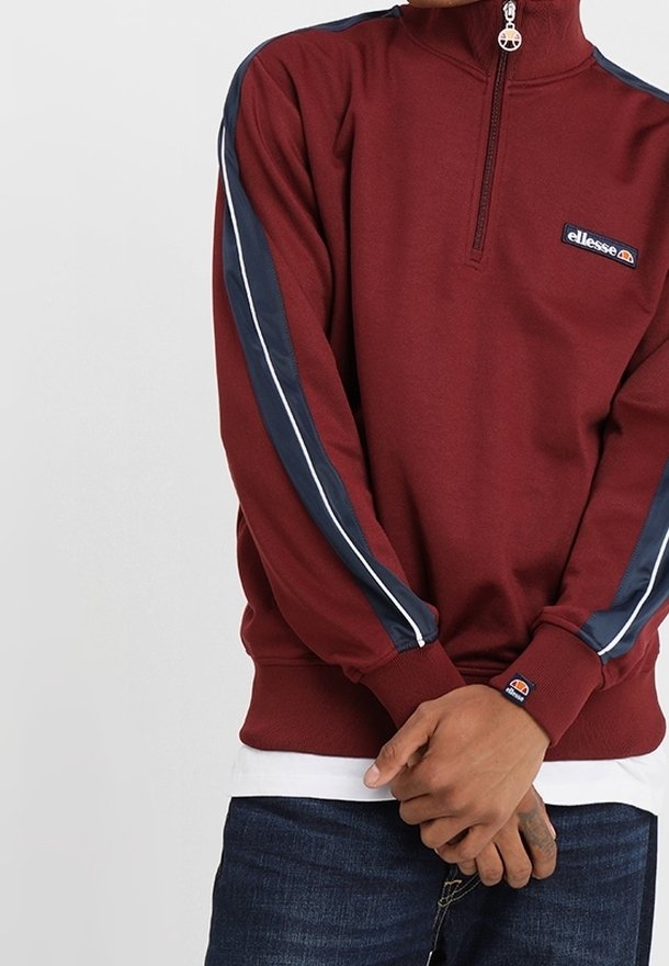 Persoon in een kastanjebruine longsleeve half-zip sweatshirt met marineblauwe en witte strepen op de mouwen, handen voor zich ineengeschoven, zichtbare blauwe jeans.