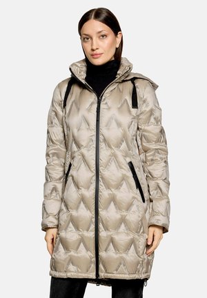 Femme portant une longue doudoune matelassée beige clair avec fermeture éclair noire et poches, par-dessus un pull à col roulé noir.