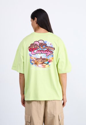 Personne aux longs cheveux foncés portant un t-shirt vert clair surdimensionné avec un motif coloré sur le thème des bonbons et un texte au dos, pantalon cargo beige.