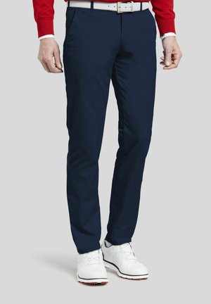 Homme portant un pantalon bleu marine, une ceinture blanche, des chaussures de golf blanches et un pull rouge, debout sur un fond gris.