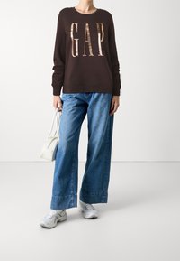 Sudadera marrón con "GAP" en letras brillantes, combinada con jeans de mezclilla azul de pierna ancha y zapatillas blancas, sosteniendo una pequeña bolsa blanca.
