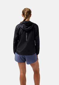 Veste légère noire avec capuche, arborant un logo réfléchissant "Brooks" au dos, associée à un short de sport bleu.