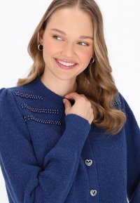 Cardigan blu navy in morbido tessuto a maglia, con dettagli di perline lungo le spalle e bottoni metallici a forma di cuore. Design a maniche lunghe.