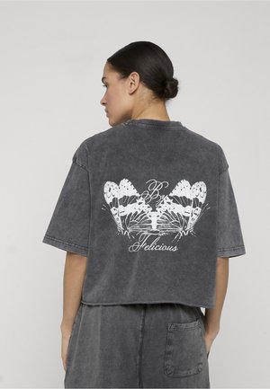 Kvinde med mørkt hår samlet i en knold iført en falmet sort oversized t-shirt med en hvid sommerfuglegrafik og teksten "By Felicious" på ryggen.