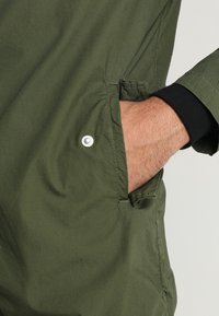 Kurtka oliwkowo-zielona z teksturowanej tkaniny. Posiada boczną kieszeń z srebrnym zatrzaskiem. Mankiety są czarne i prążkowane.