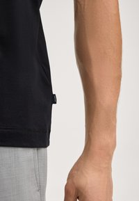 Zwarte shirt met korte mouwen en een gladde textuur, voorzien van een label aan de zijkant. Een arm zichtbaar naast lichtgrijze broek, die huidskleur en details toont.