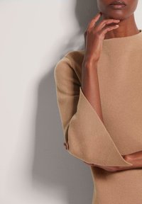 Beige Stricktop mit hohem Kragen und breiten, ausladenden Ärmeln, das aus einem strukturieren Stoff gefertigt ist. Die Hand des Modells liegt nahe am Gesicht.