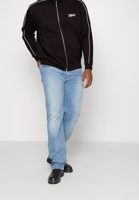 Veste noire à fermeture éclair avec des bandes blanches sur les côtés et le logo "Tigers", associée à un jean bleu clair et des sabots noirs, posant sur un fond uni.