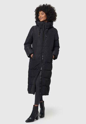 Lang zwarte pufferjas met een capuchon, rits en drukknoopsluiting; gewatteerd ontwerp; zakken aan de zijkanten; gedragen over zwarte jeans en laarzen met hak.