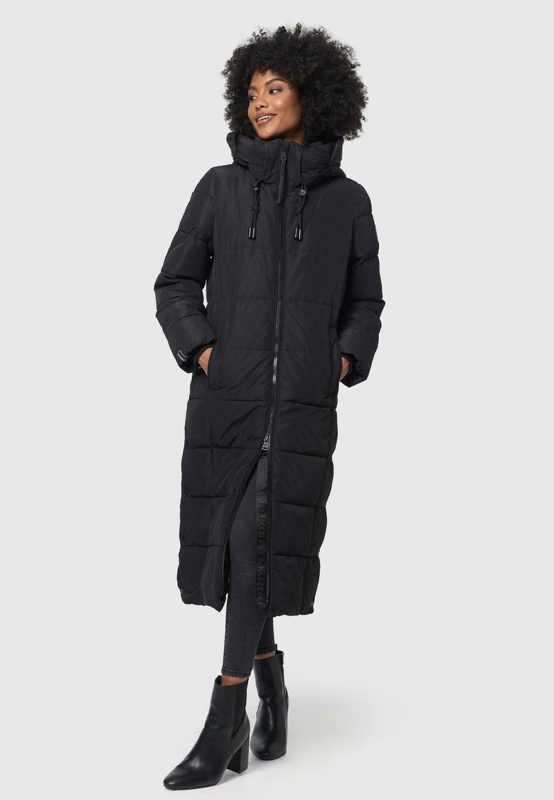 Abrigo acolchado negro largo con capucha, cremallera y cierre de botones a presión; diseño acolchado; bolsillos en los laterales; usado sobre jeans negros y botas de tacón.