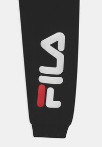 Svarta mjukisbyxor med en stor vit FILA-logotyp och röd accent, tillverkade av mjukt tyg med ribbad mudd vid ankeln.