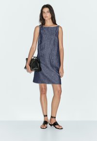 Abito senza maniche in denim, lungo fino al ginocchio, di colore blu scuro. Il modello tiene una piccola borsa nera. Indossa sandali neri flat e accessori minimi.