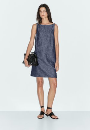 Abito senza maniche in denim, lungo fino al ginocchio, di colore blu scuro. Il modello tiene una piccola borsa nera. Indossa sandali neri flat e accessori minimi.