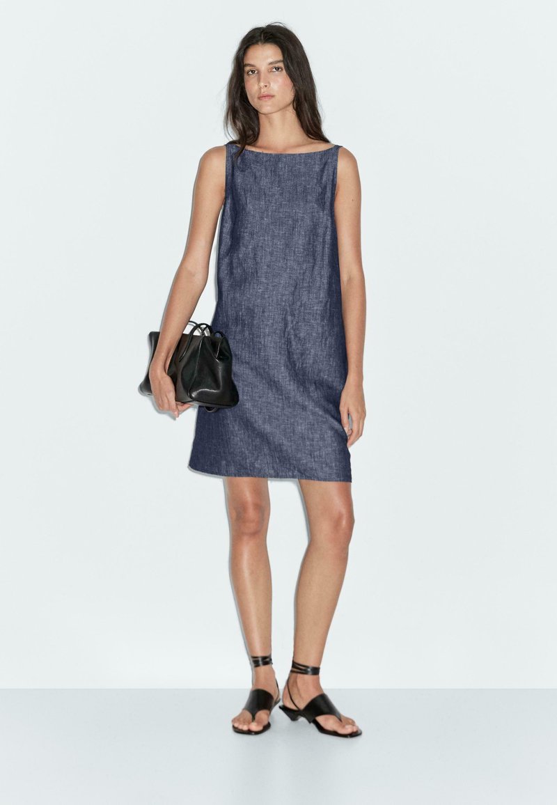 Abito senza maniche in denim, lungo fino al ginocchio, di colore blu scuro. Il modello tiene una piccola borsa nera. Indossa sandali neri flat e accessori minimi.