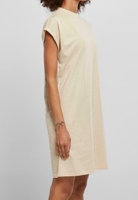 Beige puuvillamekko, jossa on lyhyet käännetyt hihat ja pyöreä kaula-aukko. Mekko on löysästi istuva ja ulottuu reiteen saakka, ja sen pinta on sileä.