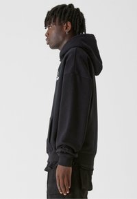 Fekete kapucnis pulóver kenguru zsebbel, bordázott mandzsettákkal és hem. Az anyag lágy és textúrált hatású, minimális logó részlettel.