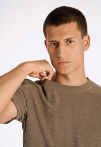 T-shirt in cotone marrone con scollatura a girocollo ribattuta, tessuto texturizzato e maniche corte, leggermente tirato indietro dalla mano di un modello.