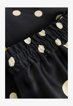 Schwarzer Stoff mit großen cremefarbenen Punkten, gerafftem elastischen Bund und glänzender, glatter Textur. Nahaufnahme der Materialdetails.