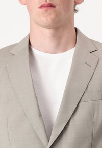 Blazer cinza claro feito de tecido texturizado, com lapelas recortadas, um pequeno bolso no peito e uma camisa branca de gola redonda por baixo.