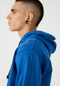 Sudadera azul con cordón, hecha de una tela suave. Presenta una capucha forrada y un ajuste relajado, mostrando el perfil lateral del usuario.