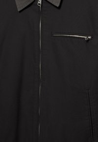 Černá bunda se středovým zipem, špičatým límcem, vodorovnou zipovou kapsou na hrudi a svislou boční kapsou z textilního materiálu.