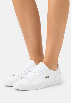 Zapatillas de cuero blanco con textura lisa, que presentan un diseño de perfil bajo y cierre de cordones. Un pequeño logo verde de Lacoste es visible en el lateral.