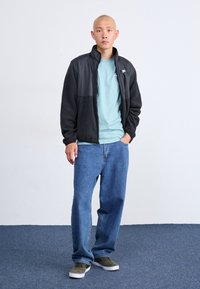 Άνδρας που στέκεται με τα χέρια στις τσέπες, φορώντας μαύρη ζακέτα, ανοιχτό μπλε μπλούζα Nike, φαρδιά μπλε τζιν και πράσινα αθλητικά παπούτσια σε μπλε χαλί.